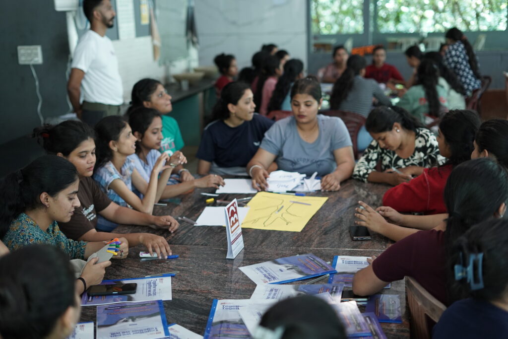 Participants in AEL T-shirts are gathered in small groups, focused and engaged in solving a hands-on challenge during an experiential learning session. They appear thoughtful, collaborative, and actively involved in discussion and strategy.