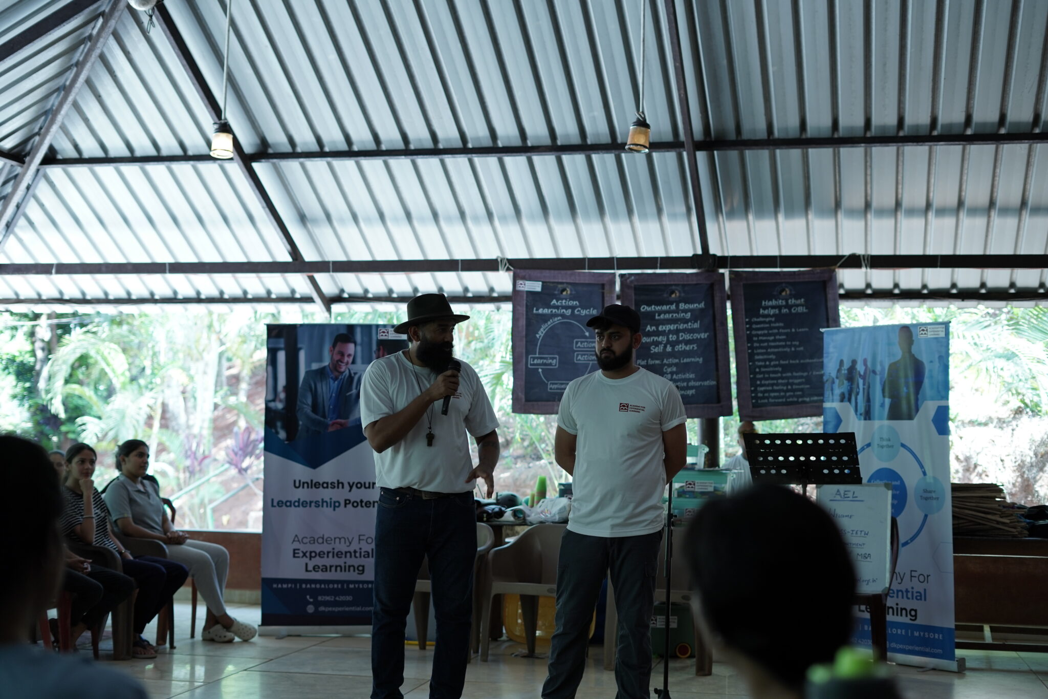 An AEL Program Director is seen speaking thoughtfully to a group of seated participants during a reflection session. The participants are listening attentively, surrounded by a calm outdoor setting that encourages introspection and dialogue.