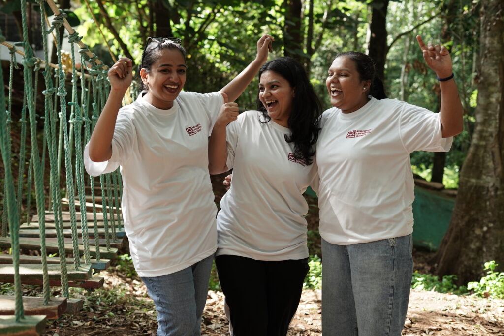 Three young women wearing AEL T-shirts are smiling brightly and posing for a photo during an experiential learning program. They look cheerful, confident, and visibly enjoying the training experience