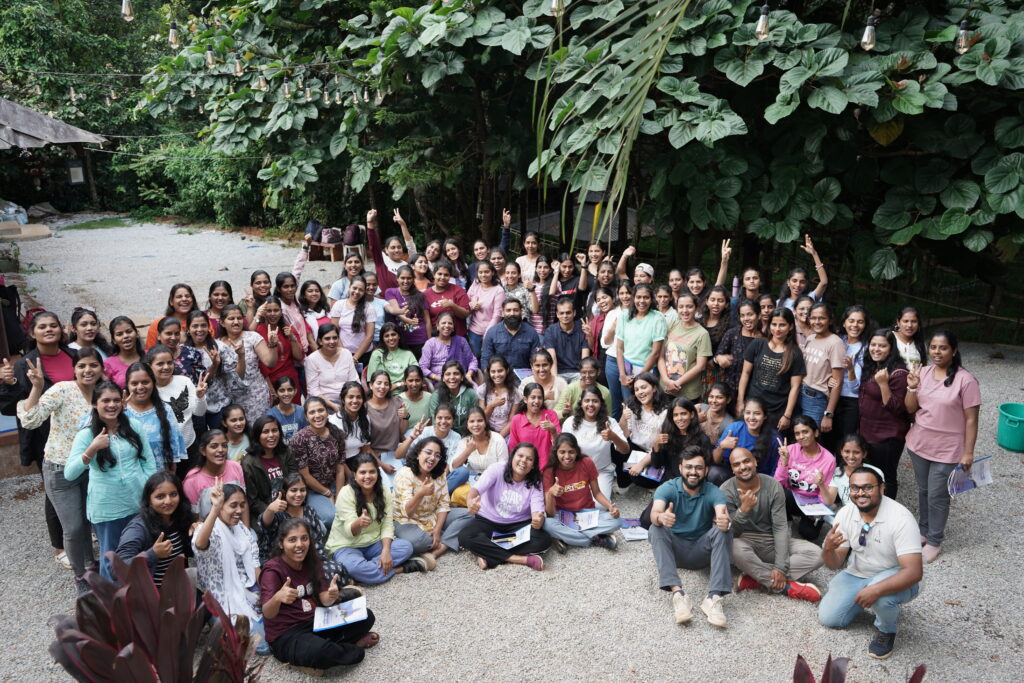 A large group of participants, all wearing AEL T-shirts, are gathered together outdoors for a group photo at the end of their experiential learning program. They are smiling, posing enthusiastically, and radiating a sense of unity and accomplishment.